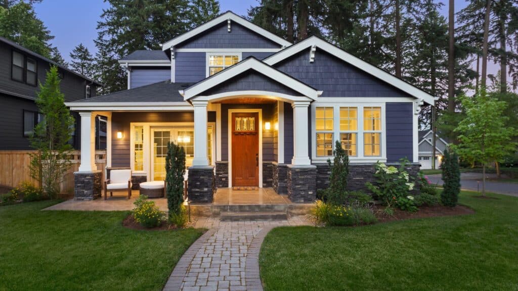 Modern blue house with white trim and stone accents, surrounded by lush greenery.