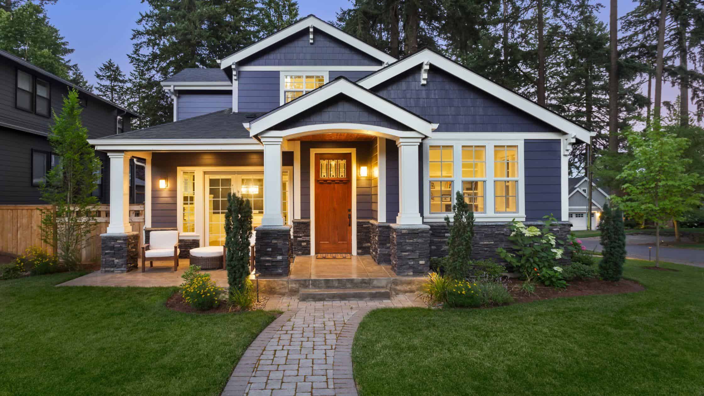 Modern blue house with white trim and stone accents, surrounded by lush greenery.