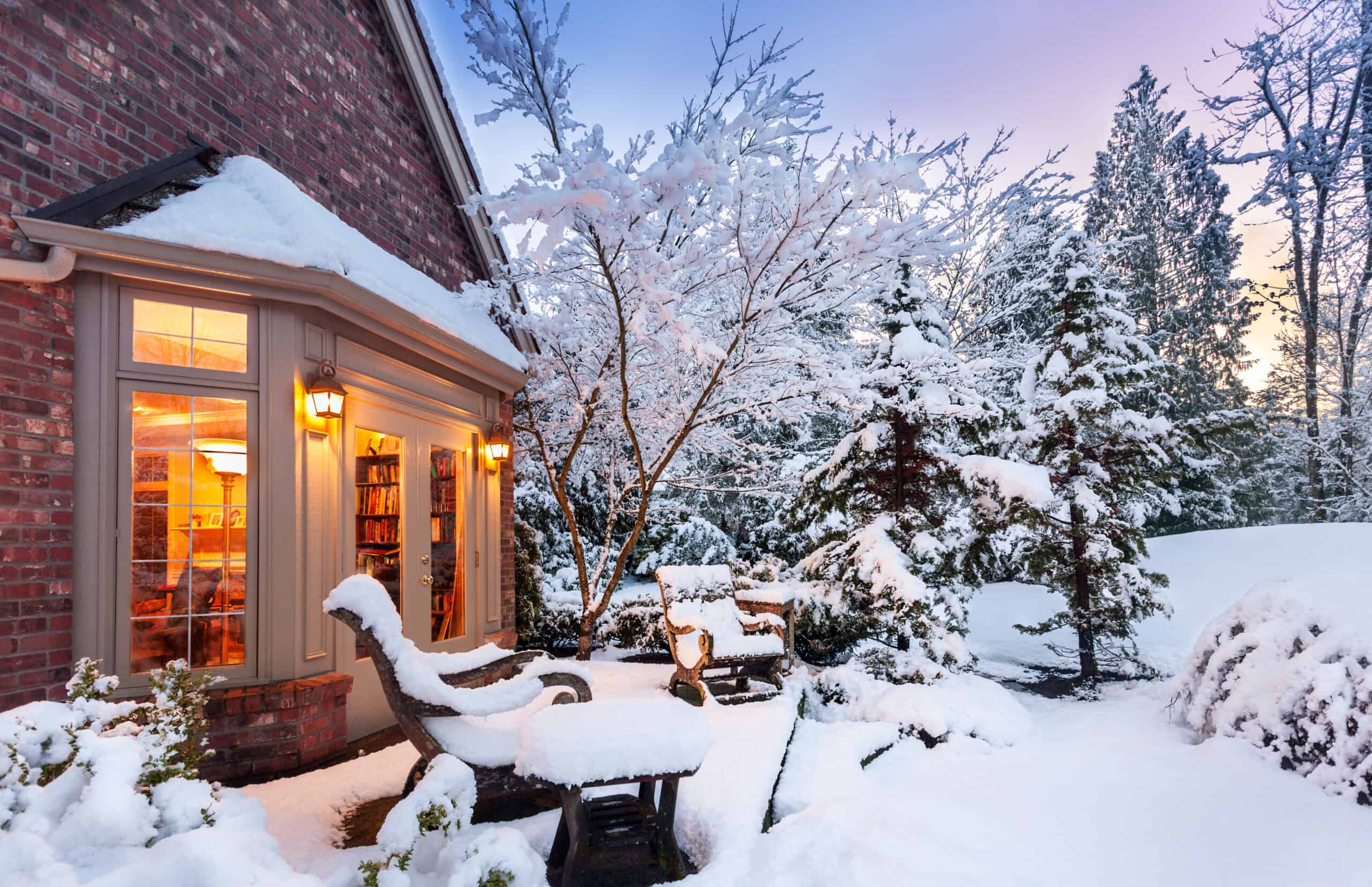 Snow-covered garden with house, trees, and outdoor furniture in winter.